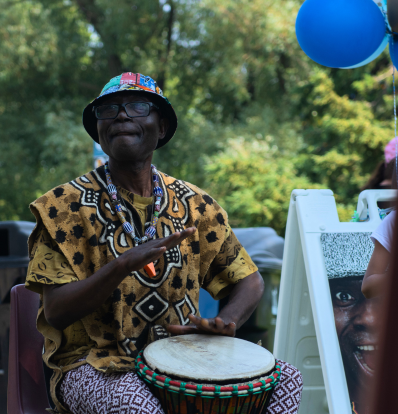 Njacko playing a djembe drum in a park. He wears a colourful shirt and bucket hat.