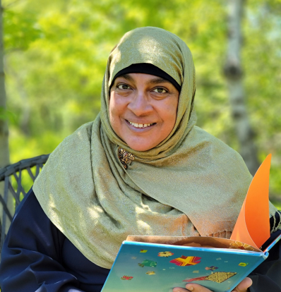 Rukhsana is pictured outside with green trees in the background. She wears a green hijab and a nice big smile. She is holding her book King for a Day