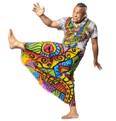 Sergio Xocolate dressed in traditional bright, multicoloured Brazilian clothing standing in a Capoeira pose; one leg up and arms out, one in front, one behind.