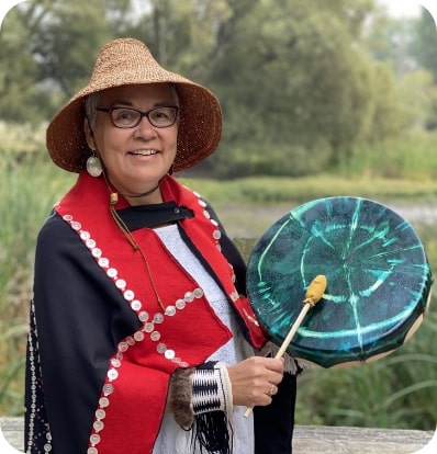 Kung Jaadee pictured outside with trees behind her. She wears her traditional Haida hat and other regalia and she's playing her green drum